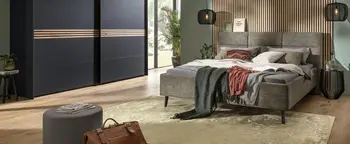 Schlafzimmer mit grauem Polsterbett, Lamellenwand hinter dem Kopfteil und dunklem Schwebetürenschrank links.